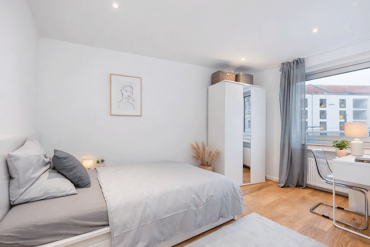 Blick in ein weißes Schlafzimmer mit großem Bett und Kleiderschrank. Rechts am Fenster steht ein Schreibtisch