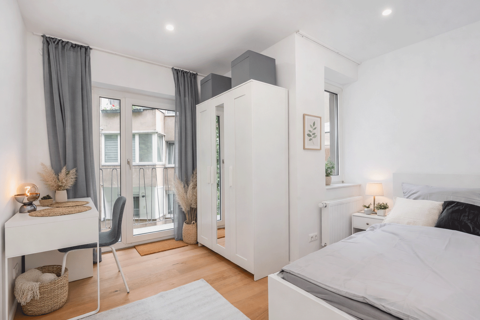 Blick in ein helles Schlafzimmer mit weißen Kleiderschrank und grauen Vorhängen. Links ist ein Schreibtisch zu sehen.